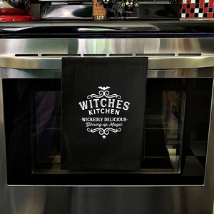 Pode incluir: Toalha de cozinha preta com texto branco que diz "Witches Kitchen - Wickedly Delicious - Stirring up Magic".