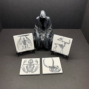 May include: Four white ceramic coasters with black and white illustrations of animal skeletons. The coasters are displayed on black stands. A black figurine of a hooded figure is in the center of the image.