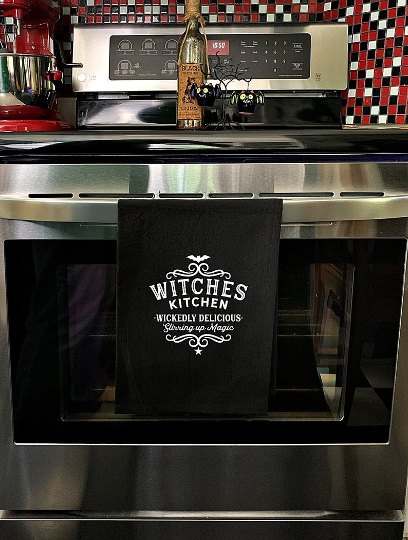 Pode incluir: Toalha de cozinha preta com texto branco que diz "Witches Kitchen - Wickedly Delicious - Stirring up Magic".