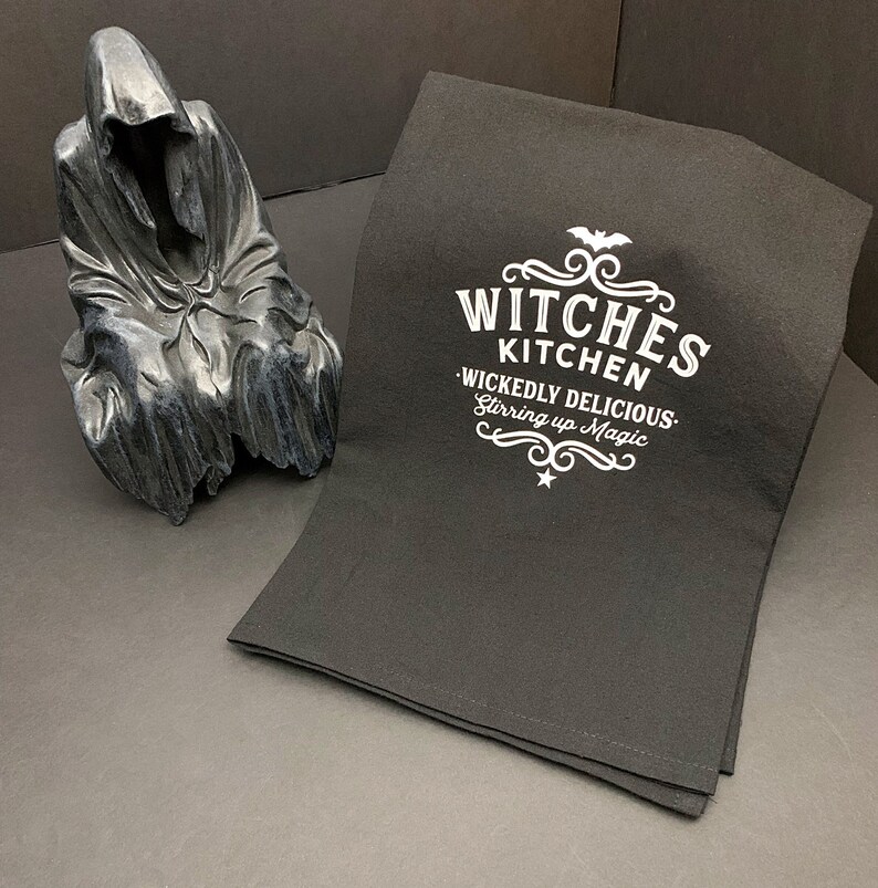 Pode incluir: Toalha de cozinha preta com texto branco que diz "Witches Kitchen - Wickedly Delicious - Stirring up Magic" com um design de morcego e estrela.