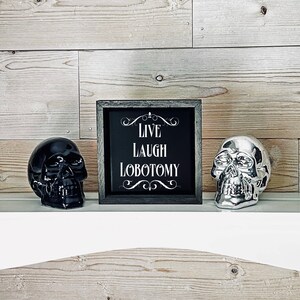 May include: A black framed sign with white text that reads "Live Laugh Lobotomy" with decorative flourishes. The sign is sitting on a white shelf with a black skull and a silver skull on either side.