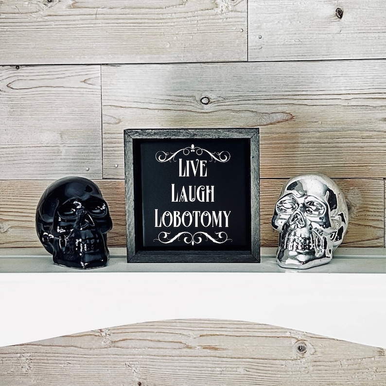 May include: A black framed sign with white text that reads "Live Laugh Lobotomy" with decorative flourishes. The sign is sitting on a white shelf with a black skull and a silver skull on either side.