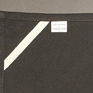 Pode incluir: Um avental de tecido preto com uma faixa diagonal branca e uma etiqueta branca que diz "100% algod&atilde;o" com instru&ccedil;&otilde;es de cuidados.