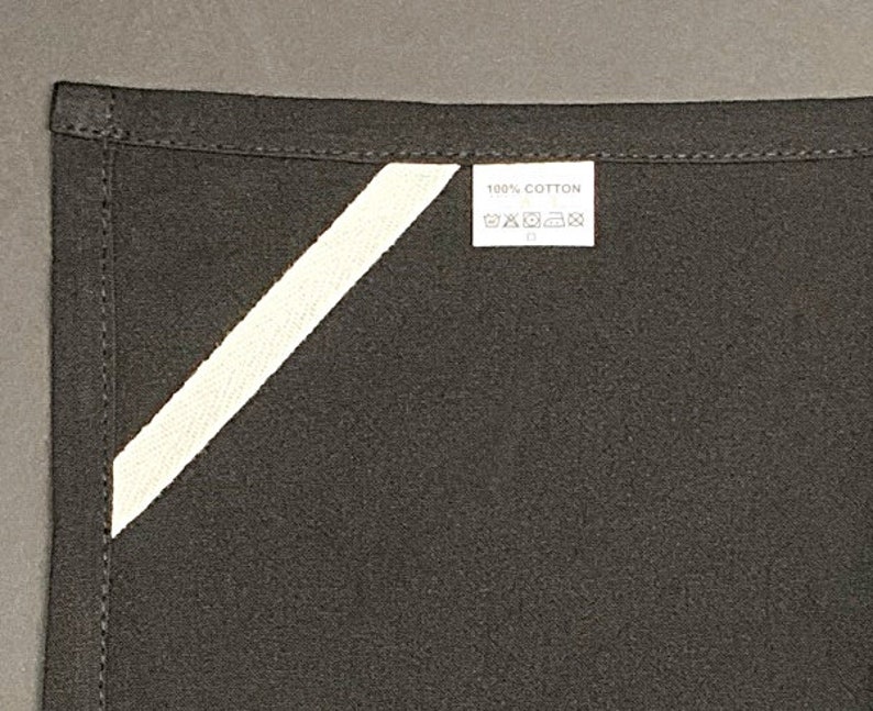 Pode incluir: Um avental de tecido preto com uma faixa diagonal branca e uma etiqueta branca que diz "100% algod&atilde;o" com instru&ccedil;&otilde;es de cuidados.