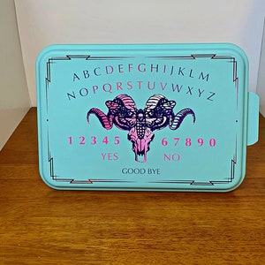 May include: A light blue rectangular cake pan with a black and white Ouija board design. The design features a skull with horns and a butterfly, the letters A-Z, the numbers 1-9 and 0, and the words "YES", "NO", and "GOOD BYE".