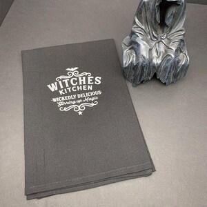 Pode incluir: Uma toalha de cozinha preta com bordado branco que diz "Witches Kitchen - Wickedly Delicious - Stirring up Magic".