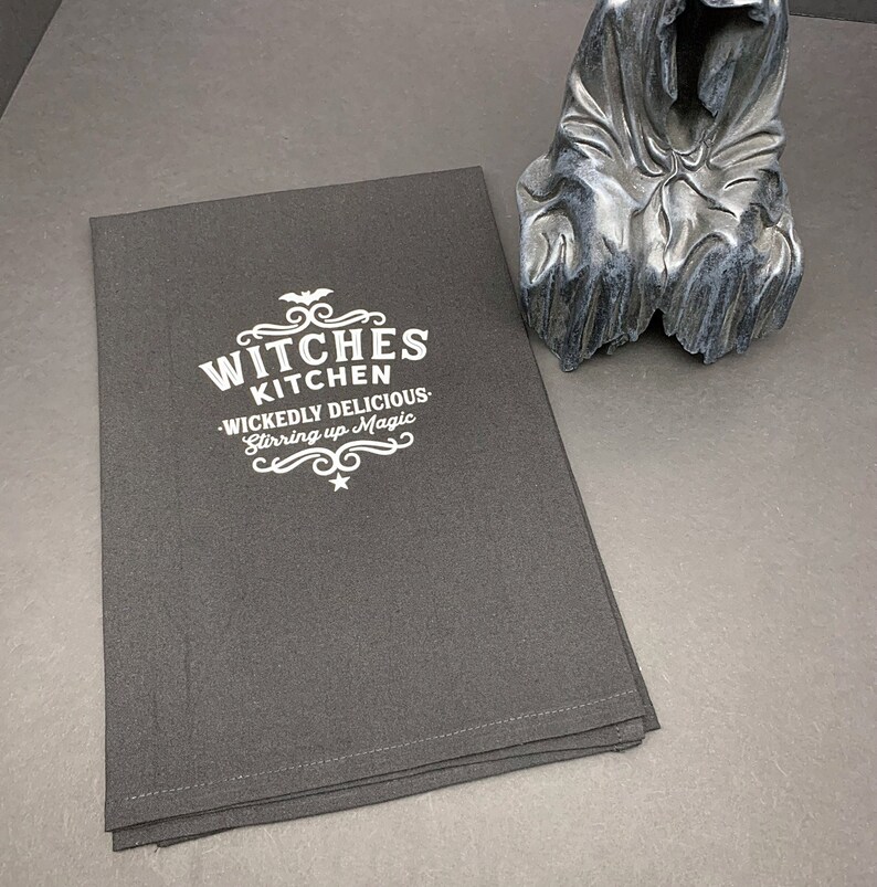 Pode incluir: Uma toalha de cozinha preta com bordado branco que diz "Witches Kitchen - Wickedly Delicious - Stirring up Magic".