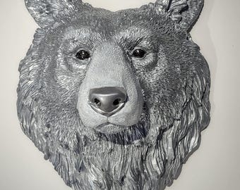 May include: A silver-colored bear head wall sculpture. The bear's fur is textured, and the nose and inner ears are a lighter shade of silver. The eyes are dark, and the sculpture is mounted on a white wall.