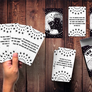 May include: A deck of black and white cards with the words "Twin Flame Truths" on the box. The cards feature black and white illustrations of a couple with stars and a crescent moon. The cards have quotes about twin flames, such as "The universe is fighting for our souls to be together. All the signs you have been noticing are not coincidences."