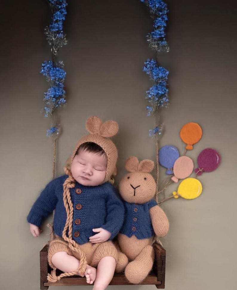 Pode incluir: Um beb&ecirc; dormindo vestindo um su&eacute;ter de tric&ocirc; azul e cal&ccedil;as de tric&ocirc; marrom senta-se em um balan&ccedil;o de madeira com um brinquedo de pel&uacute;cia marrom em forma de coelho. O balan&ccedil;o &eacute; decorado com bal&otilde;es coloridos.