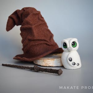 Peut inclure: Un chapeau de sorcier en feutre marron avec une pointe pointue, une figurine de hibou blanche aux yeux verts et une baguette noire sur une surface en bois. Le hibou a un motif gris et blanc sur son corps.