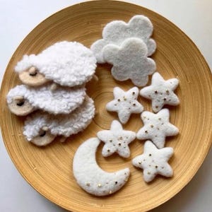 Może przedstawiać: Drewniany talerz z filcowymi dekoracjami: trzy białe owce, dwie szare chmury, pięć białych gwiazd ze złotymi akcentami i biały półksiężyc ze złotymi akcentami. Przedmioty ułożone w kapryśnym, niebiańskim motywie.