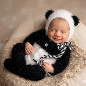 May include: A fuzzy black and white panda-themed outfit for an infant, consisting of a long-sleeved romper with white buttons and a matching bonnet featuring rounded ears. The soft, textured knit fabric covers the arms and legs completely.