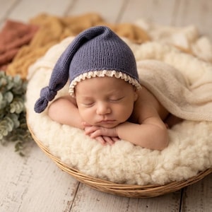 May include: A newborn baby sleeping in a woven basket lined with a cream-colored faux fur blanket. The baby wears a dark blue knit cap with a knotted tip and a delicate cream-colored ruffled trim along the edge, partially covered by a soft beige wrap.