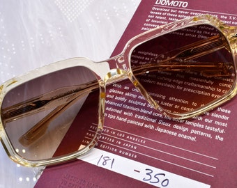May include: Clear-framed sunglasses with brown gradient lenses. The glasses rest on a burgundy card with text, including "DOMOTO" and "JACQUES MARIE MAGE." The limited edition number is 181.