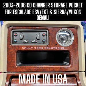 May include: A brown wood-grain CD changer storage pocket for a 2003-2006 Escalade, Sierra, or Yukon Denali. The pocket has a black interior and a silver trim. The text "VAULT-TECH SOLUTIONS" is printed on the pocket. The text "MADE IN USA" is printed on the bottom of the pocket.