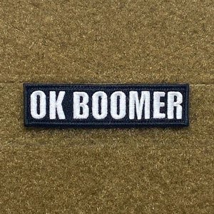 Op de afbeelding: Een zwart-witte geborduurde patch met de tekst "OK BOOMER" op een bruine achtergrond.