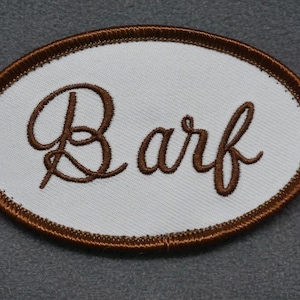 Peut inclure: Un écusson ovale brodé de couleur marron avec un tissu blanc au centre. Le mot "Barf" est brodé en lettres cursives marron.