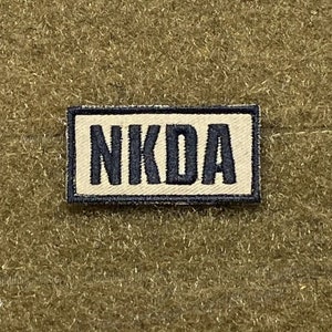 May include: A tan and black rectangular patch with the text "NKDA" in black letters.