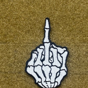 Pode incluir: Etiqueta bordada branca de um dedo médio de esqueleto, com costuras pretas, sobre um fundo de tecido verde.