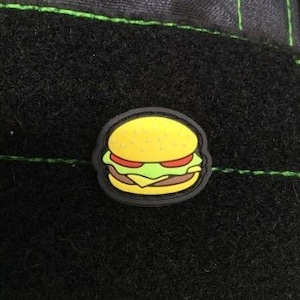 May include: A yellow, black, green, and red embroidered patch shaped like a hamburger. The patch is designed to be sewn onto clothing or bags.