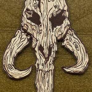 May include: Embroidered patch featuring a stylized skull with curved horns, resembling a wooden carving. The patch has a light brown and dark brown color scheme, with detailed stitching that mimics wood grain. The patch is on a green background.