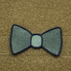 May include: A teal bow tie patch with a navy blue border. The bow tie is centered on a textured, tan background. The patch has a woven texture.