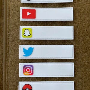Könnte beinhalten: Eine Anordnung weißer rechteckiger Aufnäher mit Logos von Social-Media-Plattformen wie TikTok, YouTube, Snapchat, Twitter, Instagram und einem Pokéball. Sie sind vertikal auf einem braunen Stoffhintergrund angeordnet.