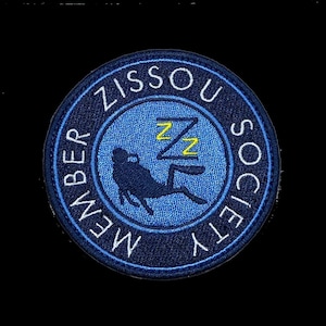 Puede incluir: Un parche bordado azul y negro con el texto "Zissou Society Member" en blanco y un buceador durmiendo estilizado con "zz" amarillas.