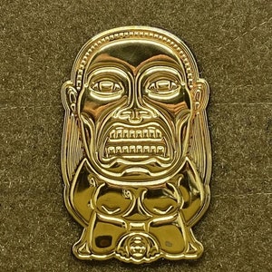 May include: Gold-coloured metal pin featuring a detailed depiction of a tiki mask with a wide, open mouth and a menacing expression.