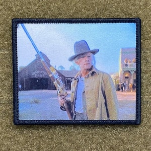 May include: A vintage-style embroidered patch featuring a man in a cowboy hat holding a rifle, standing in front of a wooden building. The patch has a black border and a white background.