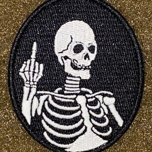 Puede incluir: Un parche bordado en blanco y negro de un esqueleto haciendo el dedo medio. El esqueleto es blanco con contornos negros y está sobre un fondo circular negro.