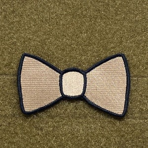 Puede incluir: Un parche de pajarita beige con costuras negras, perfecto para añadir un toque de fantasía a una bolsa o chaqueta.