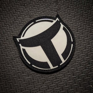 May include: A black and white circular patch with a stylized letter "T" in the center. The patch has a white background and a black border.