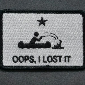 Pode incluir: Um remendo branco bordado com contorno preto. O remendo apresenta um desenho animado de uma pessoa em uma canoa, com uma estrela acima da canoa. O texto "OOPS, I LOST IT" está abaixo da canoa.
