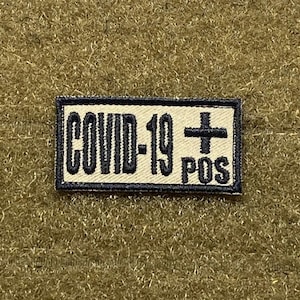 Puede incluir: Un parche beige con costuras negras y el texto "COVID-19+ POS" en negro.
