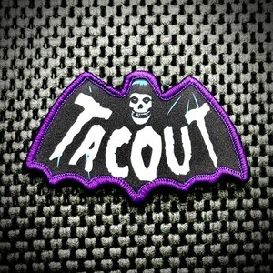 May include: A black and white embroidered patch with a bat design and the word "TACOUT" in white lettering. The patch has a purple border.