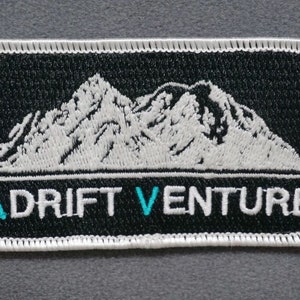 May include: White embroidered mountain range design on a black patch with white stitching around the edges. The text "ADrift VENTURE" is embroidered in white with a turquoise "V".