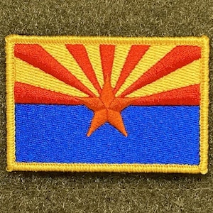 May include: An embroidered patch featuring the state flag of Arizona. The flag depicts a red star in the centre of a yellow sun with red rays, all on a blue background.