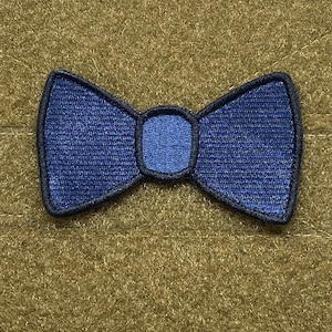 May include: A blue bow tie patch with a black outline. The patch is made of fabric and has a textured surface.