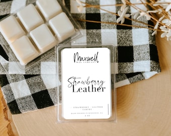 May include: A clear plastic container holding six wax melts, next to a container with a label that reads "Maxwell Wax Company Strawberry Leather". The background includes a black and white checkered cloth and wooden surface.