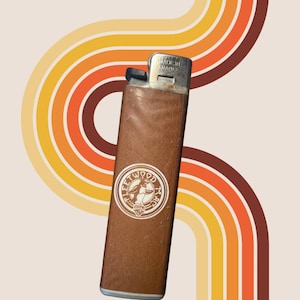 May include: A vintage brown leather lighter with a silver metal top. The lighter has a white circular logo with text and an illustration. The lighter is set against a retro background with curved stripes in shades of orange, yellow, and brown. The text "MADE IN FRANCE" is visible.