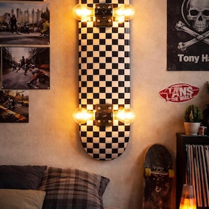 May include: A skateboard repurposed as a wall-mounted light fixture. The board features a black and white checkerboard pattern and is fitted with two Edison-style light bulbs. The fixture is mounted on a neutral-colored wall, creating a unique decor piece.