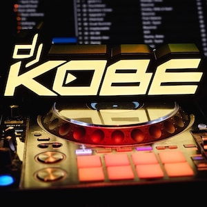 May include: A DJ setup with the name "KOBE" illuminated in white against a black background. The DJ controller features a silver turntable, red lights, and orange pads. The overall theme is music and entertainment.
