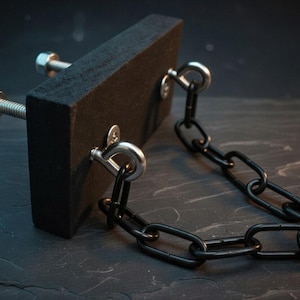May include: A black rectangular block with two bolts and a black chain attached. The bolts are silver and the chain is made of black metal links. The block appears to be made of a dense, dark material.