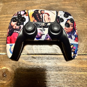 May include: A black and white PlayStation 5 controller with a custom design featuring various images of people. The controller has black handles and buttons, with a colorful design on the touchpad area.