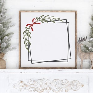 May include: A white wooden mantel with a decorative carved design. A framed print with a green and red floral wreath and a black geometric frame is centered on the mantel. Two small artificial snow-covered pine trees are on either side of the print. A small wooden reindeer figurine is on the right side of the print.