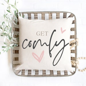 May include: A white cushion with the text "GET comfy" in black script with pink hearts. The cushion is in a woven basket with wooden beads.
