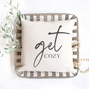 Puede incluir: Una almohada blanca con texto negro que dice "get cozy" en una fuente de escritura decorativa. La almohada está en una cesta tejida con cuentas de madera.