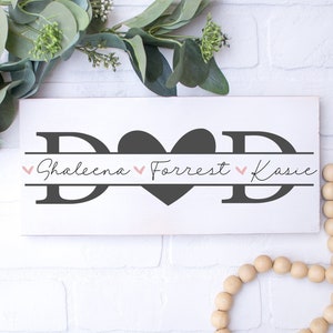 Puede incluir: Un letrero de madera blanco con la palabra "DAD" en letras negras. Un gran corazón negro se encuentra entre las letras. Los nombres "Shaleena", "Forrest" y "Kasie" están escritos en cursiva debajo de las letras.